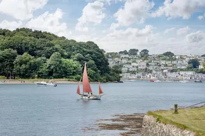 a boat with a red sail on a river with a city at Lavender Cottage in Porthscatho