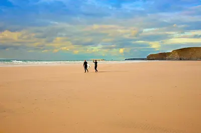 two people walking on a beach with a surfboard at Tregurrian Villas in Newquay